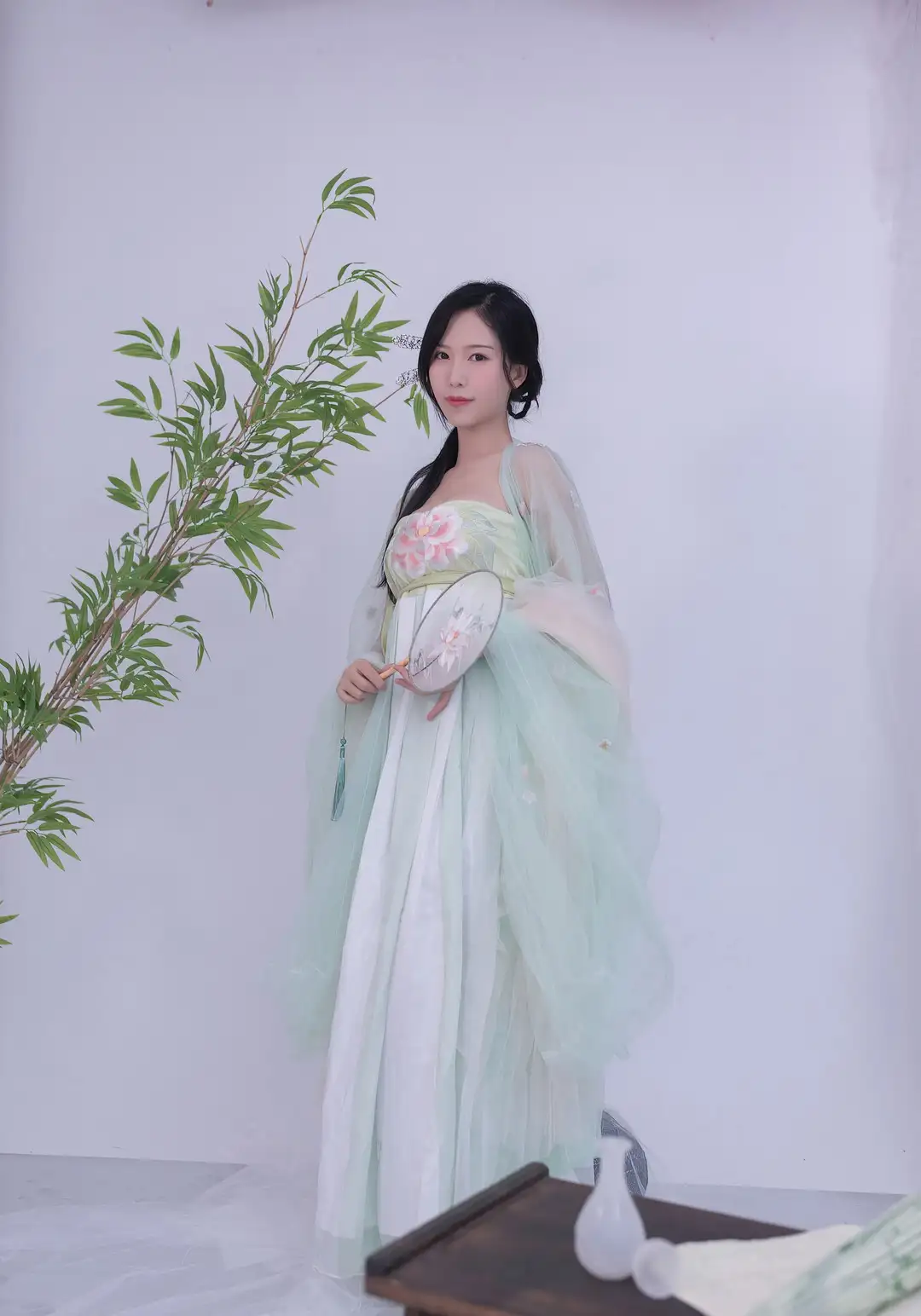 抖娘利世 淡绿齐胸襦裙 外罩薄纱大袖衫,手持团扇立于竹枝旁白色背景 抖娘利世 淡绿齐胸襦裙 外罩薄纱大袖衫,手持团扇立于竹枝旁白色背景